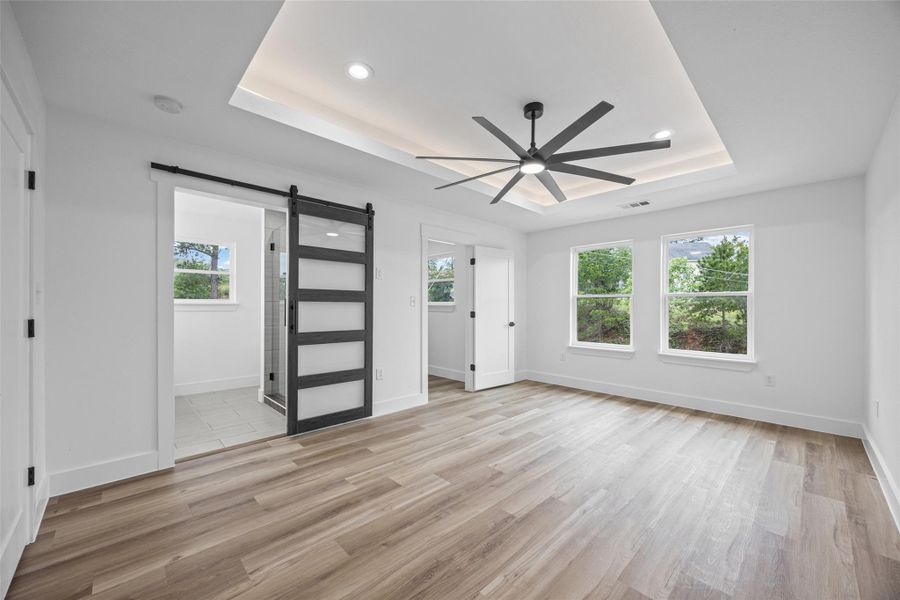 Spacious primary bedroom featuring wood-finish flooring, a contemporary ceiling fan, and a tray ceiling with recessed lighting Spacious primary bedroom featuring wood-finish flooring, a contemporary ceiling fan, and a tray ceiling with recessed lighting