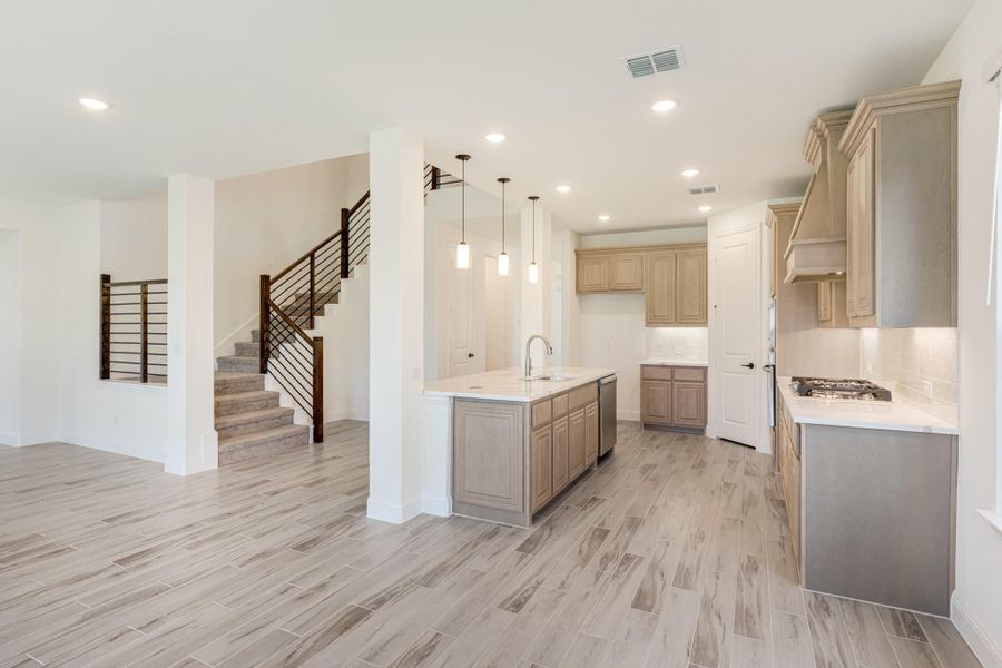 Open kitchen with center island, light wood cabinets, pendant lights, and staircase visible in background Open kitchen with center island, light wood cabinets, pendant lights, and staircase visible in background