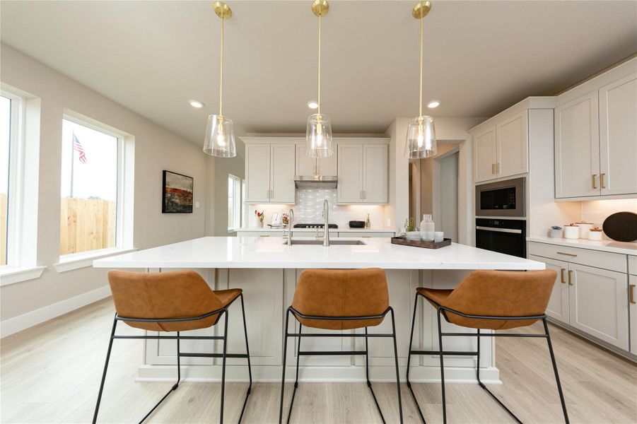 This gourmet kitchen serves as the ultimate heart of the home, showcasing a massive central island with a built-in sink and premium countertops. Bright white cabinetry lines the walls, offering abundant storage space, complemented by a built-in microwave, wall oven, and a sleek range hood over the stove. A row of elegant pendant lights illuminates the breakfast bar area, while large side windows fill the entire space with rich natural light.