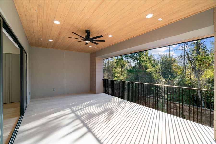 This photo showcases a spacious covered patio with a wooden ceiling and ceiling fan, offering a serene view of lush greenery through a railing. Ideal for outdoor relaxation and gatherings.