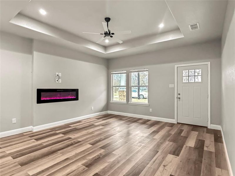 Unfurnished living room with ceiling fan, a raised ceiling, a glass covered fireplace, wood finished floors, and recessed lighting