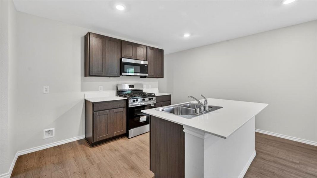 Kitchen featuring wood-finish flooring, dark wood cabinetry, white countertops, stainless steel appliances, and a central island with an integrated sink