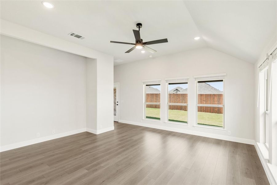 Empty living room with vaulted ceiling, ceiling fan, hardwood floors, and three windows overlooking backyard Empty living room with vaulted ceiling, ceiling fan, hardwood floors, and three windows overlooking backyard