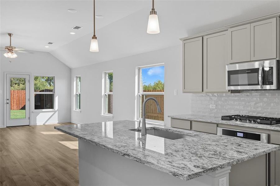 Kitchen with light stone counters, backsplash, stainless steel appliances, decorative light fixtures, and a kitchen island with sink Kitchen with light stone counters, backsplash, stainless steel appliances, decorative light fixtures, and a kitchen island with sink