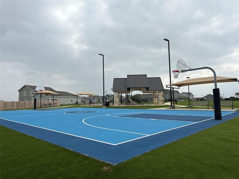Outdoor recreational space featuring a blue-surfaced basketball court, artificial turf perimeter, covered seating areas, modern streetlights, and a central pavilion with stone accents