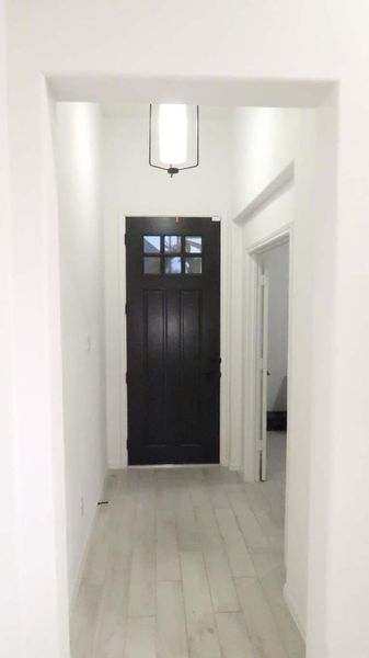 This photo shows a bright, narrow hallway with light-colored flooring, leading to a black front door. It features a modern hanging light fixture and doors to adjacent rooms on the right. This photo shows a bright, narrow hallway with light-colored flooring, leading to a black front door. It features a modern hanging light fixture and doors to adjacent rooms on the right.