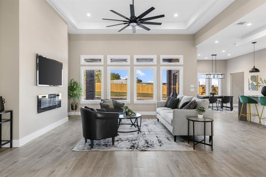 Living room featuring ceiling fan with notable chandelier, light wood-type flooring, a raised ceiling, and a wealth of natural light