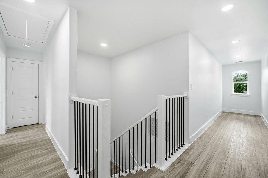 Upper-level hallway featuring wood-finish flooring, recessed lighting, a window with exterior views, and a staircase with a black metal spindle railing Upper-level hallway featuring wood-finish flooring, recessed lighting, a window with exterior views, and a staircase with a black metal spindle railing