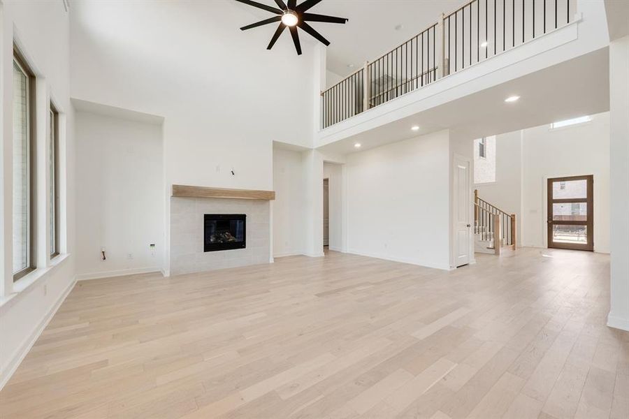 Unfurnished living room featuring a towering ceiling, light wood-style flooring, a tiled fireplace, ceiling fan, and stairs Unfurnished living room featuring a towering ceiling, light wood-style flooring, a tiled fireplace, ceiling fan, and stairs