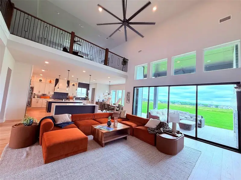 Spacious living area featuring a soaring vaulted ceiling with a large ceiling fan, a second-story interior balcony with black metal railings, and a wall of floor-to-ceiling sliding glass doors