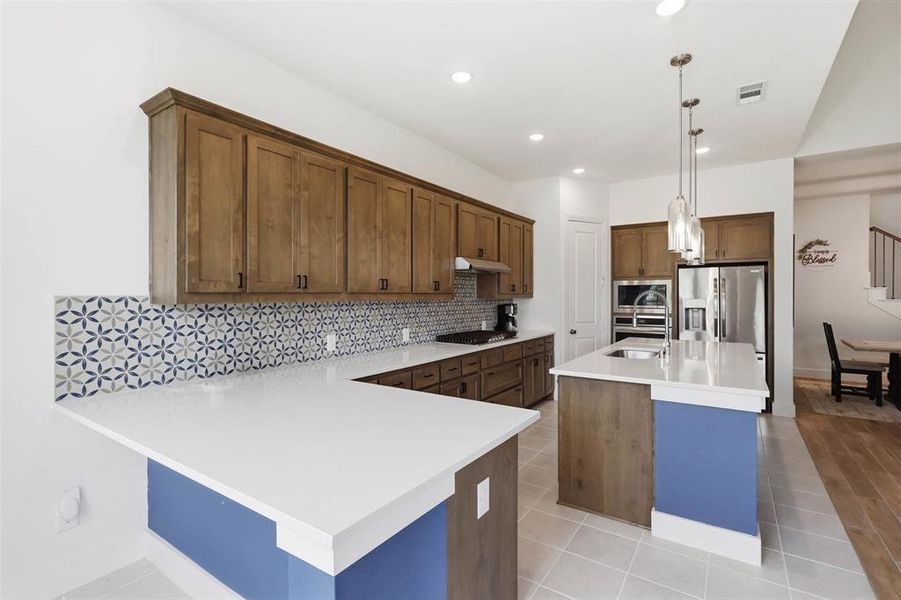 Open-concept kitchen featuring light wood-finish cabinetry, bright white countertops, a patterned tile backsplash, a central island with sink, and recessed lighting