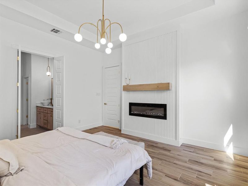 Bedroom featuring wood finished floors, a large fireplace, ensuite bath, a chandelier, and a tray ceiling
