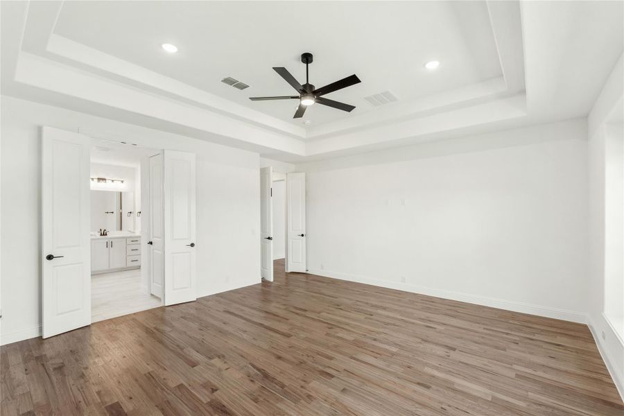 Spacious bedroom with tray ceiling, ceiling fan, hardwood floors, and ensuite bathroom visible through open door