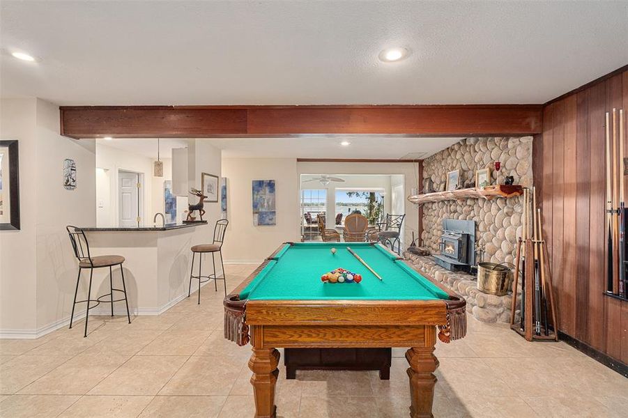Entertainment room featuring a stone fireplace with wood-burning insert, wood-finish wall paneling, a wet bar with a dark countertop, and tiled flooring