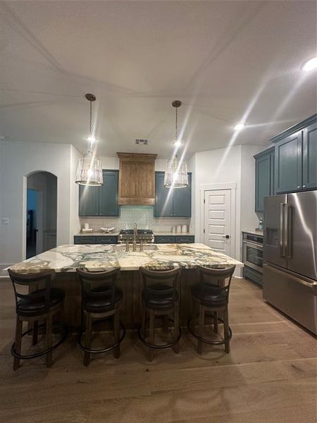 Spacious kitchen featuring a large island with seating, natural wood-finish flooring, teal cabinetry, stainless steel appliances, and pendant lighting