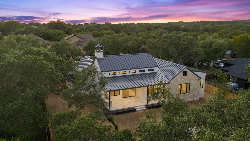 Mature trees surround a modern home with a metal roof and stone facade under twilight skies.
