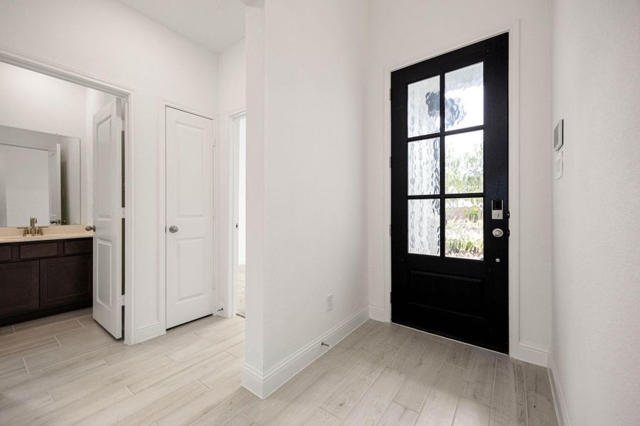 Inviting entryway with a stylish black front door featuring frosted glass panels, complemented by light wood flooring and access to adjacent rooms, including a bathroom with a modern vanity.