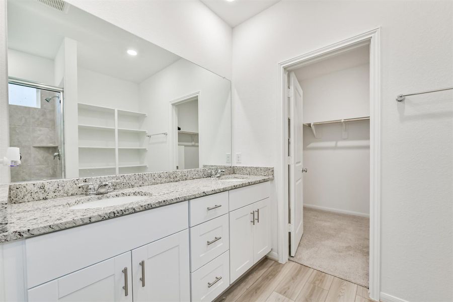 This bright, modern bathroom features double sinks with granite countertops, ample storage, and a large mirror. It offers a walk-in shower and connects to a spacious walk-in closet with shelving and hanging space.