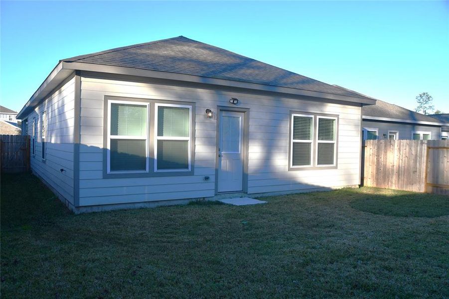 This is a photo of a single-story home with a light gray exterior and a fenced backyard. It features multiple energy-efficient windows and a central back door, situated on a grassy lawn.