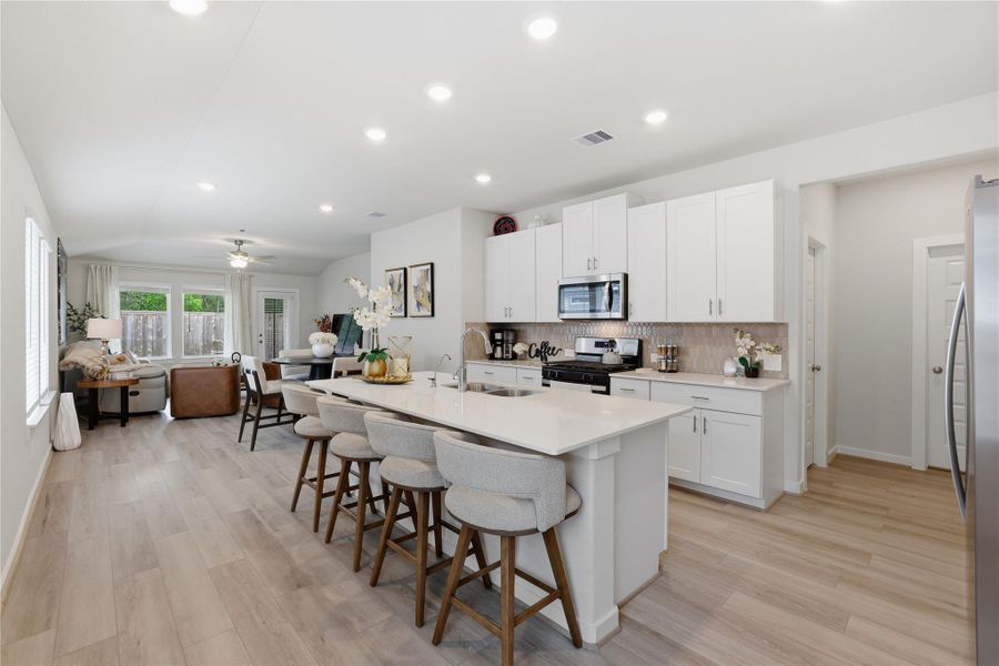 This modern open-concept kitchen and living area features sleek white cabinetry, a central island with seating, stainless steel appliances, and light wood flooring. The living space is bright, with large windows and a cozy seating area, creating an inviting atmosphere.