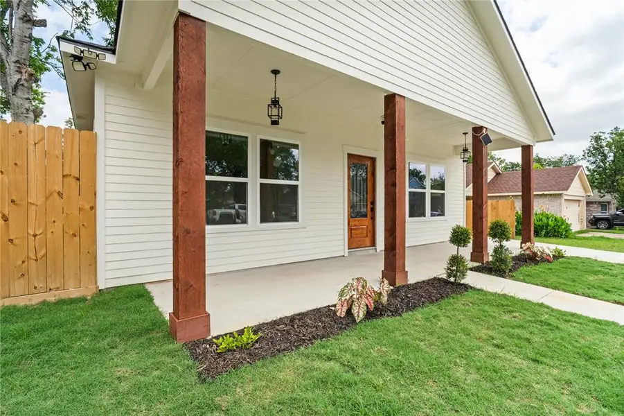 White horizontal siding exterior with a covered porch featuring stained wood columns, a paneled wood entry door with decorative glass, and a concrete walkway bordered by landscaping