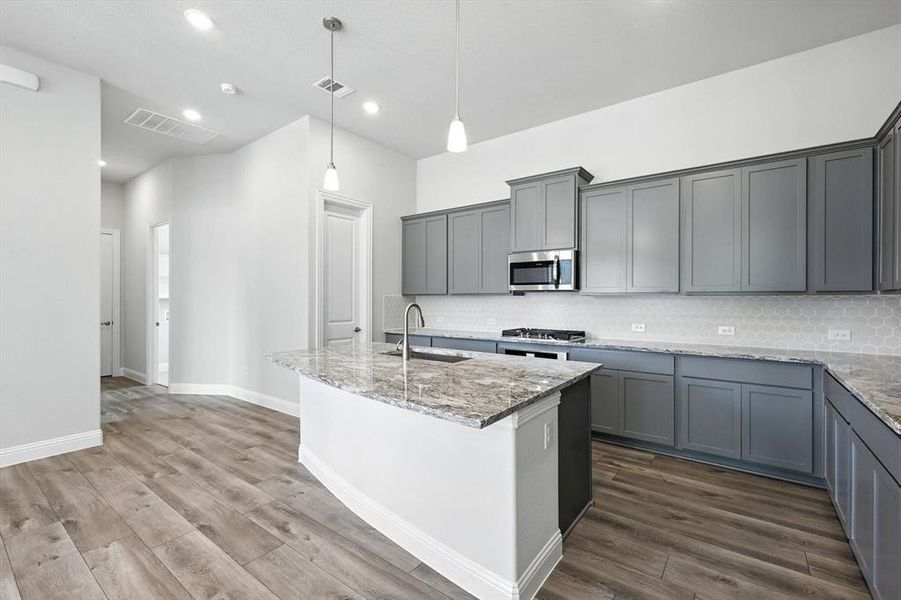 Kitchen with backsplash, light stone counters, dark wood-style flooring, a kitchen island with sink, and gray cabinetry Kitchen with backsplash, light stone counters, dark wood-style flooring, a kitchen island with sink, and gray cabinetry