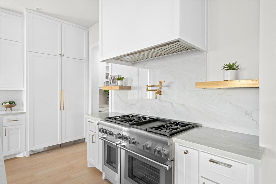 Showcasing the chef's kitchen with a professional grade double oven and range, complemented by a sleek marble backsplash, pot filler, and custom cabinetry with integrated refrigerator panels. Brass accents and floating shelves complete the modern yet timeless design. Showcasing the chef's kitchen with a professional grade double oven and range, complemented by a sleek marble backsplash, pot filler, and custom cabinetry with integrated refrigerator panels. Brass accents and floating shelves complete the modern yet timeless design.