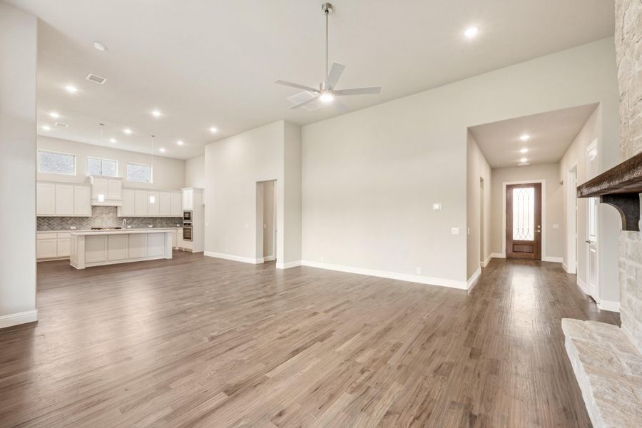 Open-concept living area with hardwood floors, high ceilings, ceiling fan, stone fireplace, and white kitchen in background.
