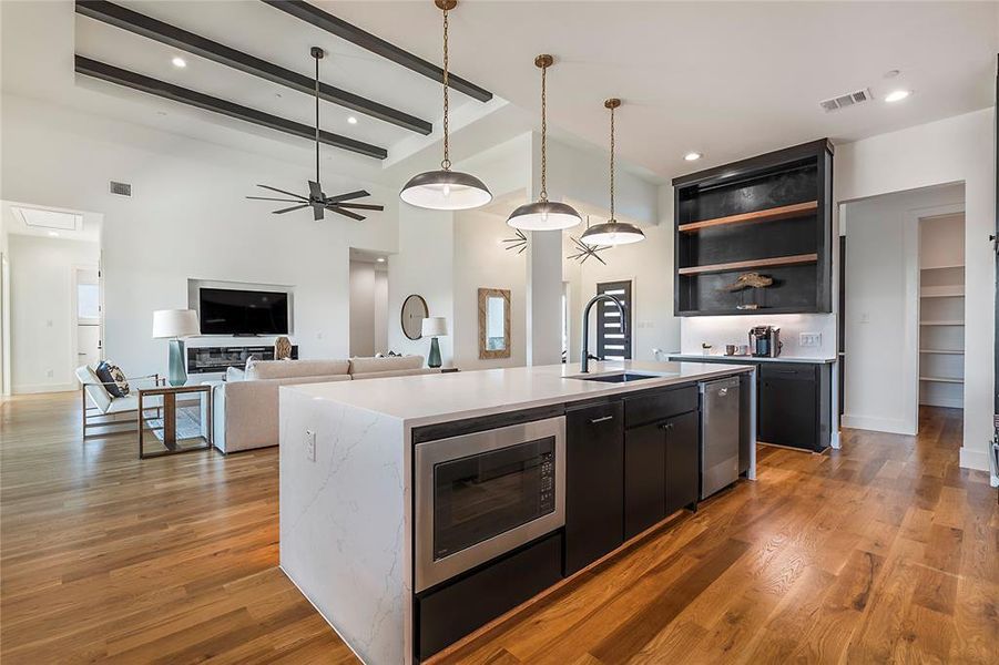 Open-concept living space featuring a spacious kitchen island with integrated microwave, wood-finish flooring, vaulted ceilings with dark accent beams, a modern ceiling fan, and recessed shelving Open-concept living space featuring a spacious kitchen island with integrated microwave, wood-finish flooring, vaulted ceilings with dark accent beams, a modern ceiling fan, and recessed shelving