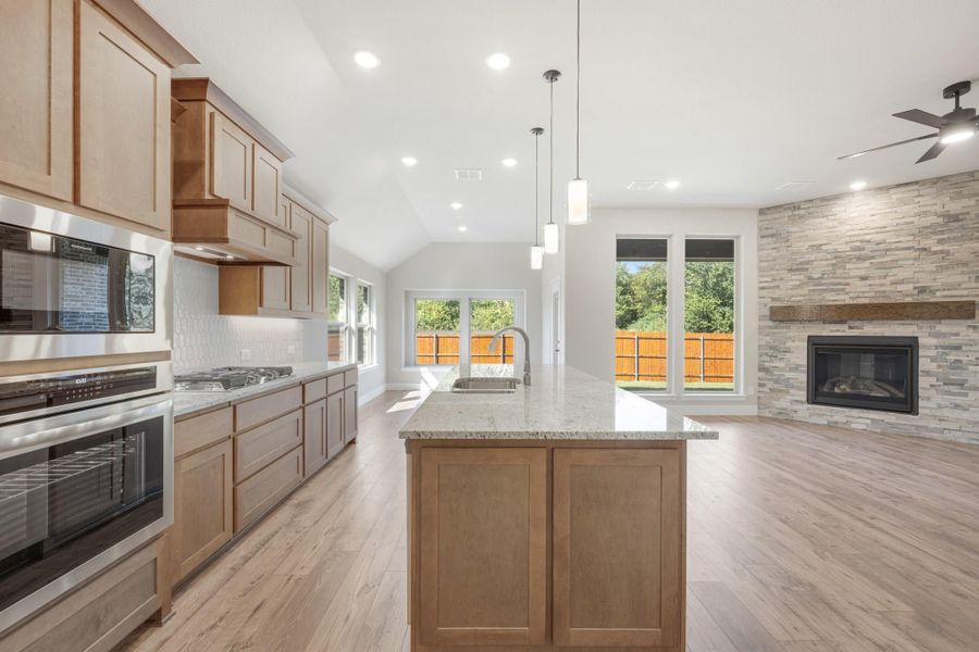 Open kitchen with wood cabinets, granite island, and stone fireplace visible in adjoining living area