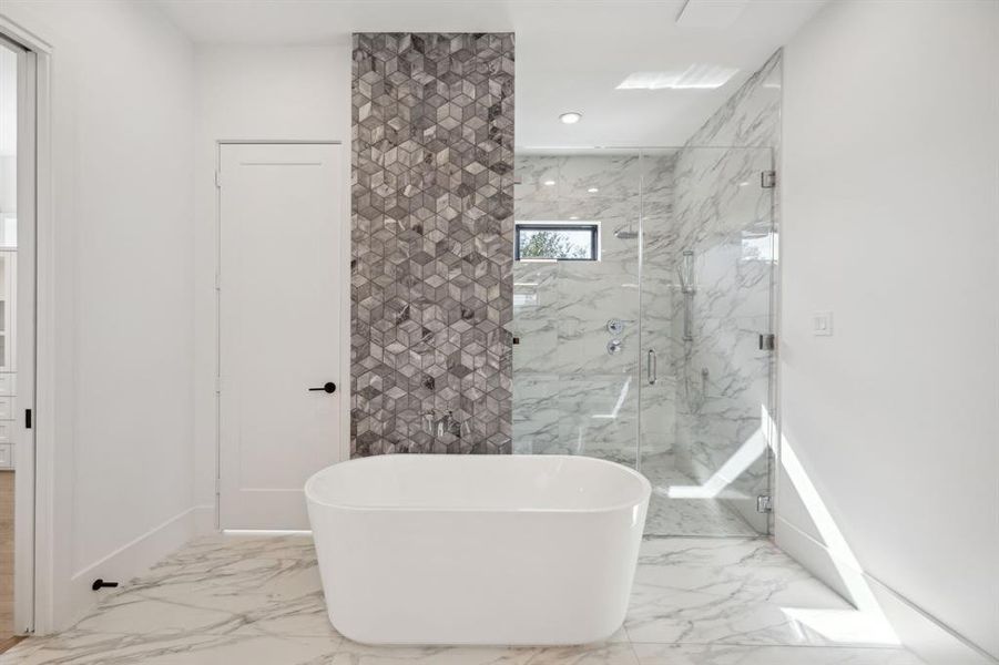 The freestanding white bathtub is positioned in front of a feature wall adorned with gray geometric marble.The bathroom features large white Italian tiles with subtle gray veining on the floor and walls of the shower.