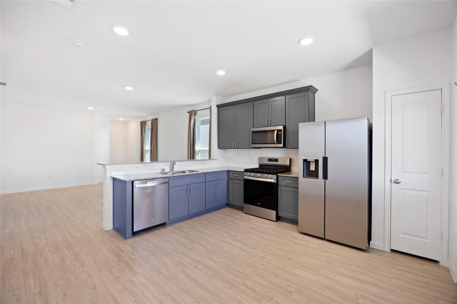 modern kitchen with sleek stainless steel appliances, including a refrigerator, oven, and dishwasher. It features stylish gray cabinetry, a spacious countertop, and an open layout that flows into the living area with light wood flooring.