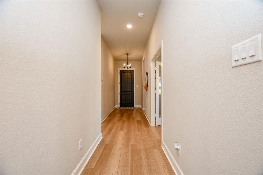 This photo shows a bright hallway with light wood flooring, neutral walls, and a ceiling light fixture. It leads to a dark door at the end, with a side room visible on the right.