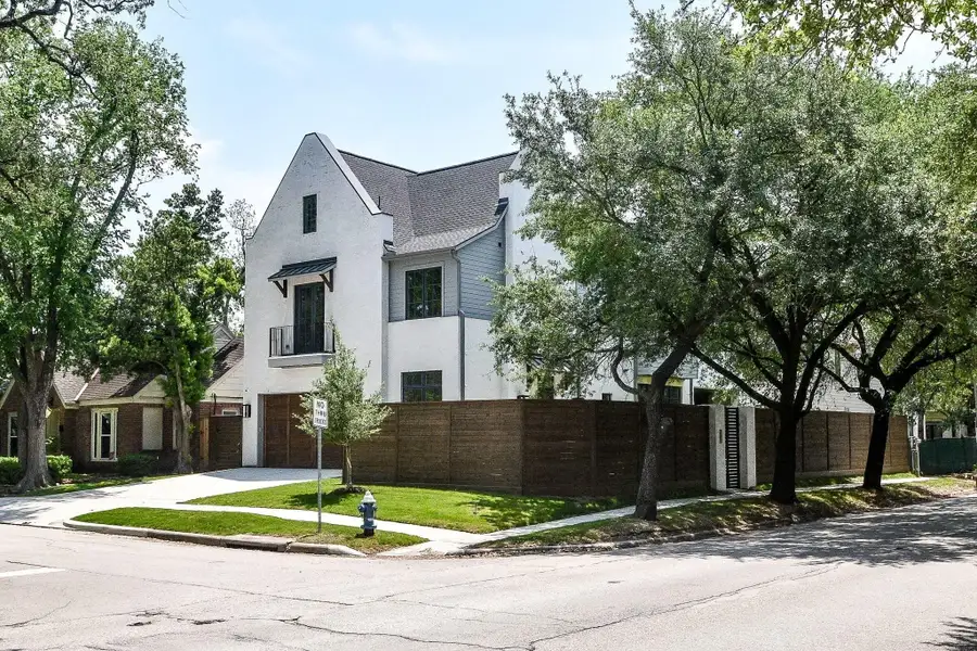 This is a modern three-story house situated on a corner lot. It features a neatly manicured lawn, large windows, and a wooden fence for privacy, surrounded by mature trees for added shade.