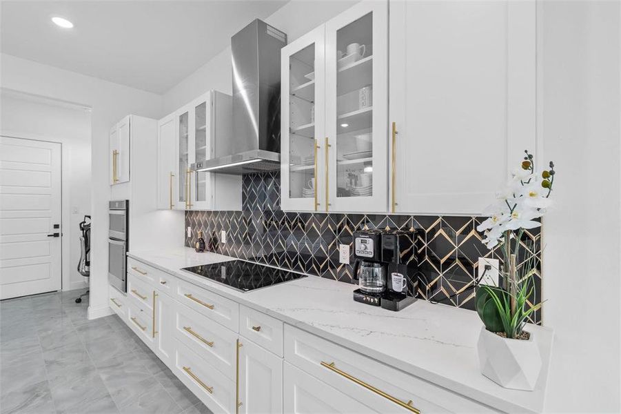 Kitchen featuring backsplash, wall chimney exhaust hood, white cabinetry, light stone countertops, and glass insert cabinets