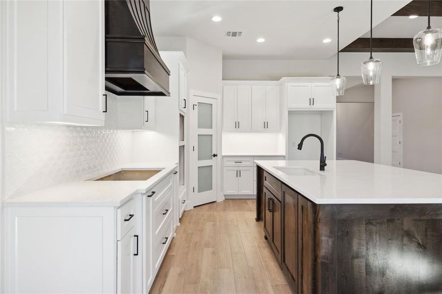 Kitchen featuring custom exhaust hood, stovetop, decorative light fixtures, white cabinets, and an island with sink