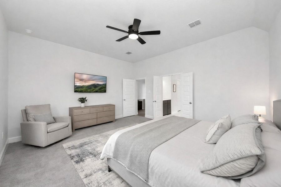 A spacious bedroom with soft, neutral tones, a generous bed, and a cozy seating area. The room includes a ceiling fan, dresser, and wall-mounted TV, with an en-suite bathroom accessed through double doors for privacy and convenience.