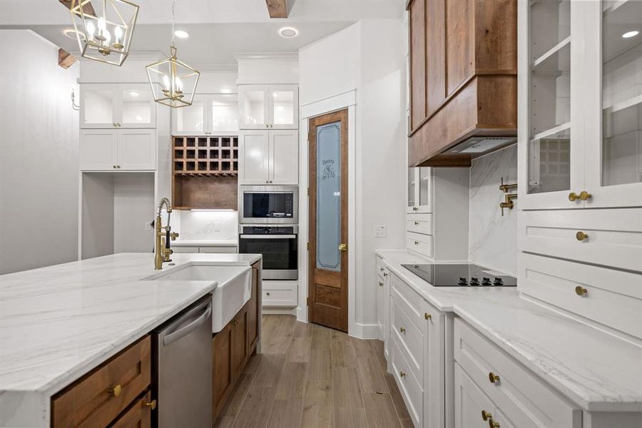 Gourmet kitchen featuring a large central island with a farmhouse sink, white perimeter cabinetry, integrated stainless steel appliances, a built-in wine rack, and a wood-finish range hood Gourmet kitchen featuring a large central island with a farmhouse sink, white perimeter cabinetry, integrated stainless steel appliances, a built-in wine rack, and a wood-finish range hood