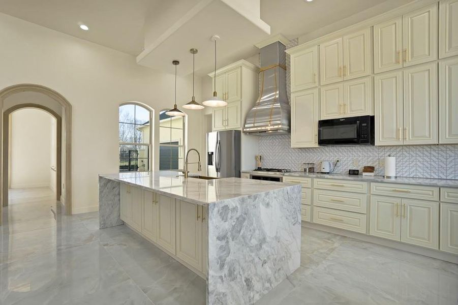 Kitchen featuring wall chimney exhaust hood, light stone counters, stainless steel fridge with ice dispenser, a center island with sink, and pendant lighting