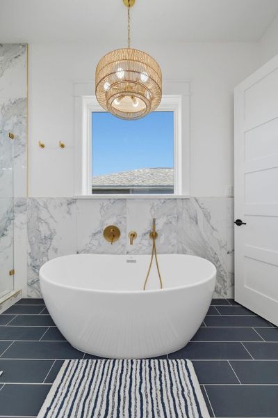 This bathroom features a modern design with a freestanding white tub against elegant marble walls. It has sleek black tile flooring, a stylish striped rug, and a chic woven chandelier. The gold fixtures add a touch of luxury, complemented by natural light from the window.