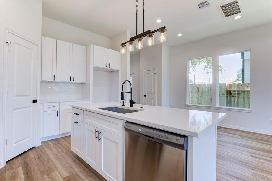 Light-filled kitchen with a generous quartz island, single-basin sink, and matte black fixtures. Crisp white cabinetry and a subway tile backsplash keep it fresh, while pendant lighting adds warmth. A window brings in natural light, and the door to the left opens to a spacious pantry.