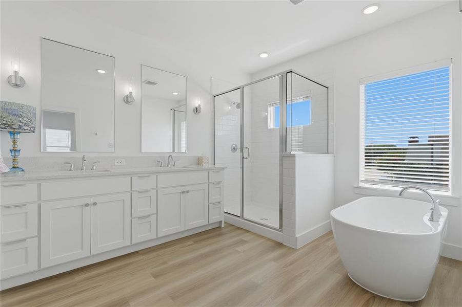 Spacious bathroom featuring a freestanding soaking tub, a frameless glass shower enclosure, a dual vanity with white cabinetry, large rectangular mirrors, and wood-finish flooring