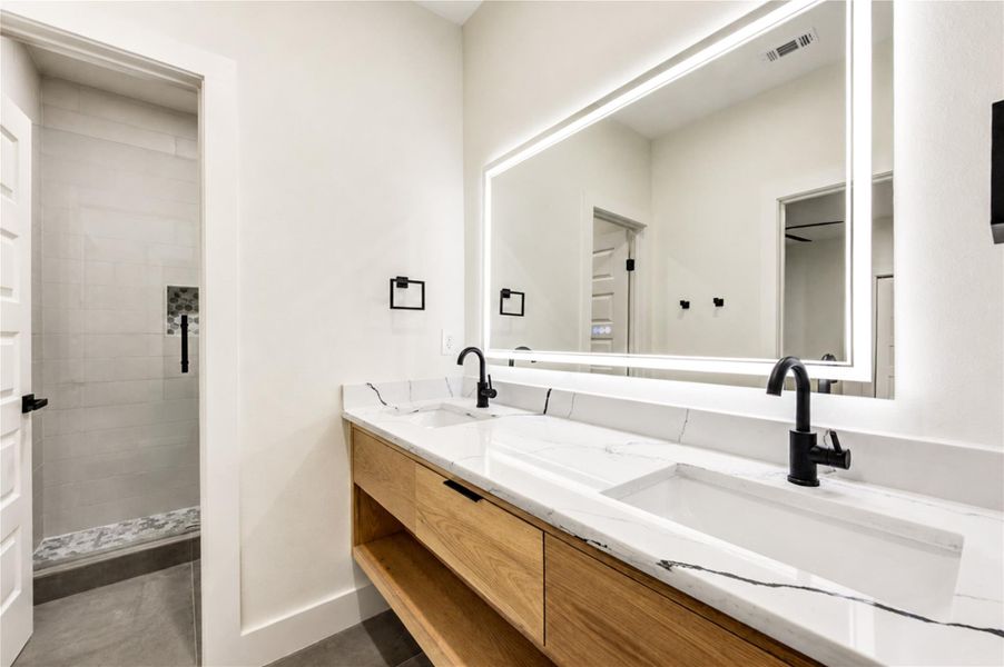 Jack and Jill bathroom featuring a dual-sink vanity with elegant black fixtures, a large illuminated mirror, and a spacious walk-in shower with modern tilework.