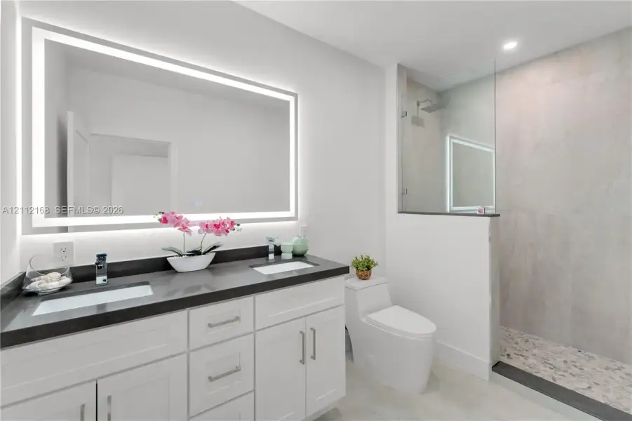 Double vanity with dark countertops, integrated rectangular sinks, wall-to-wall LED backlit mirror, walk-in glass shower with stone-style tile surround, and tile flooring Double vanity with dark countertops, integrated rectangular sinks, wall-to-wall LED backlit mirror, walk-in glass shower with stone-style tile surround, and tile flooring