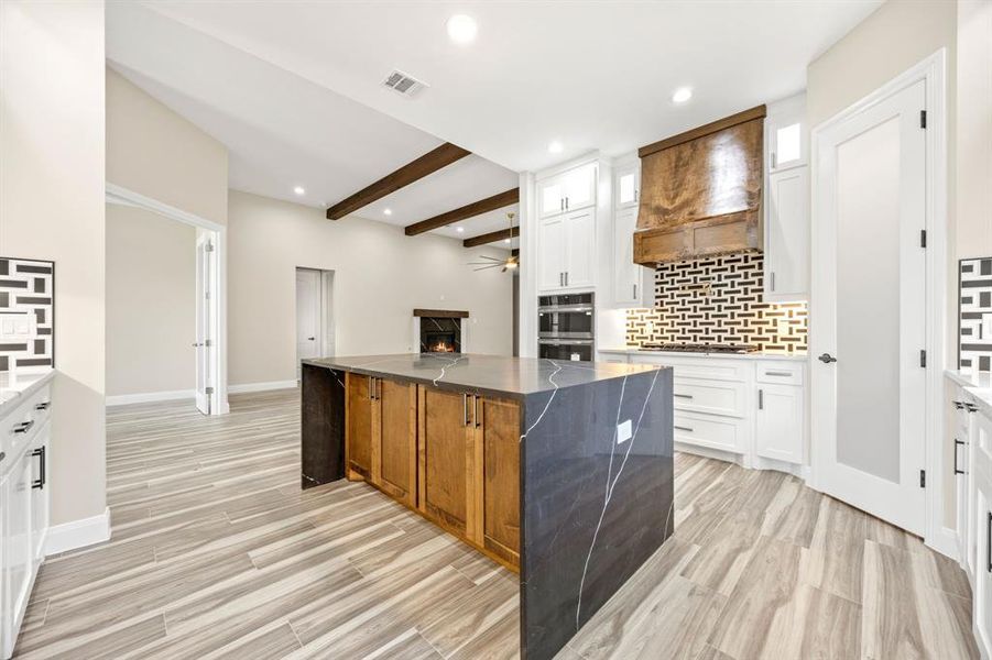 Kitchen with backsplash, custom exhaust hood, white cabinetry, beamed ceiling, and a center island