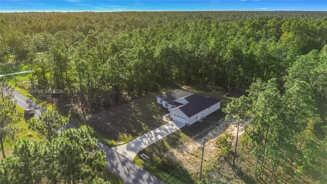 Single-story residence featuring a dark shingled roof, light-toned siding, attached garage, and a paved concrete driveway situated on a wooded lot