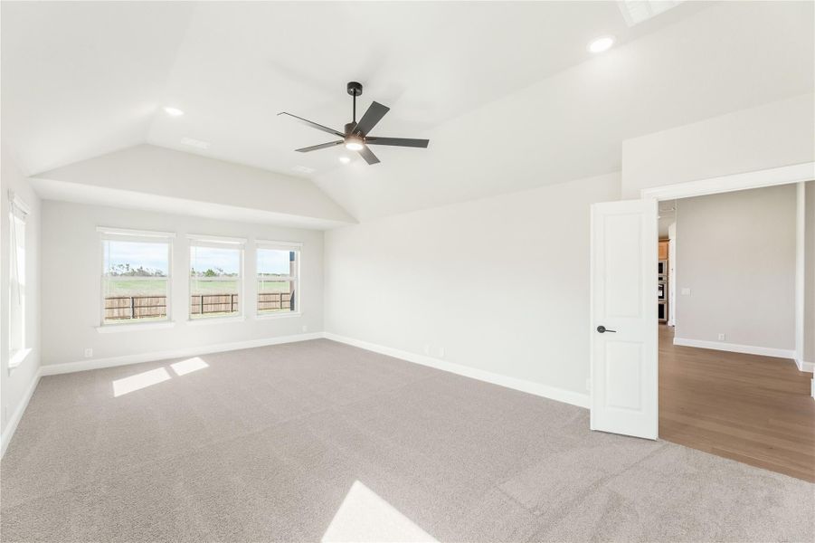 Spacious bedroom with vaulted ceiling, ceiling fan, gray carpet, and triple windows overlooking a yard Spacious bedroom with vaulted ceiling, ceiling fan, gray carpet, and triple windows overlooking a yard
