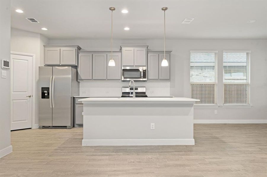 Kitchen with appliances with stainless steel finishes, gray cabinetry, hanging light fixtures, a kitchen island with sink, and light wood-type flooring