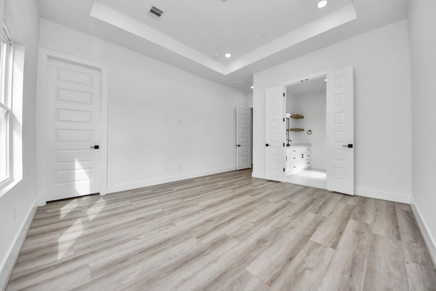 This bright, spacious room features modern wood flooring, recessed lighting, and a stylish tray ceiling. It includes a walk-in closet with double doors and has ample natural light from large windows.
