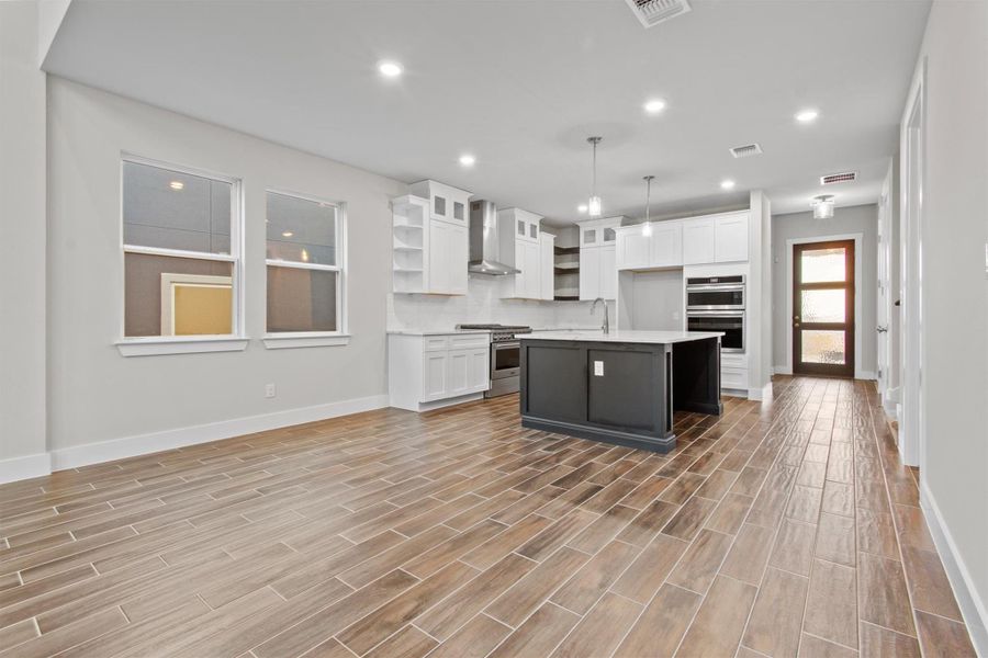 Kitchen with pendant lighting, white cabinets, wall chimney range hood, stainless steel appliances, and an island with sink